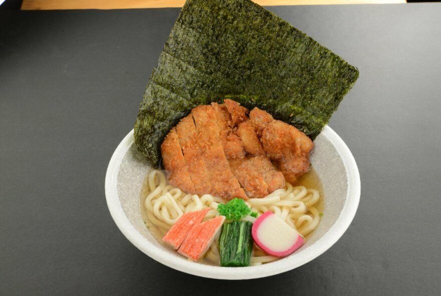 A delicious Japanese meal in a white take-away bowl with noodles, crumbed chicken and a nori sheet.