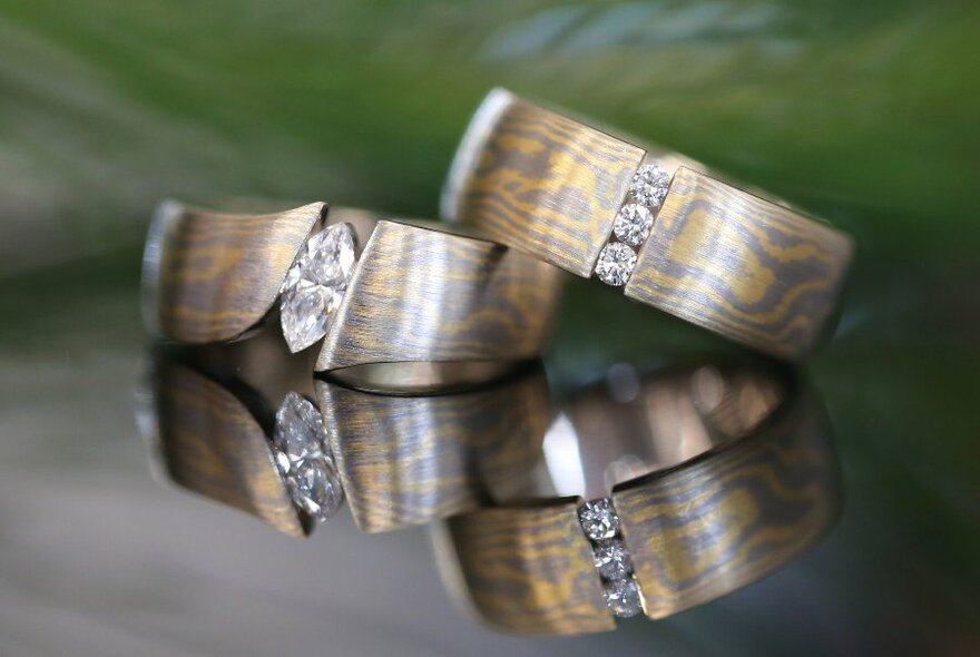 Two thick Mokume Gane diamond rings on a reflective surface.