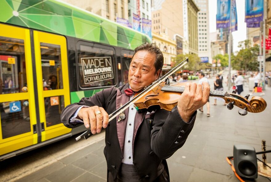 A man plays a violin in a public space while a tram goes past in the background.