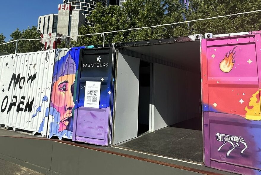 Converted shipping containers showing an entrance and painted space-themed designs on exterior, with city buildings, trees and blue sky visible behind.