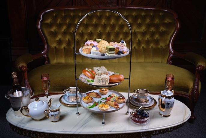 A lavish High Tea spread on a three-tiered plate, a pot of tea, cups and saucers arranged on a low marble oval table, in front of a velvet couch.