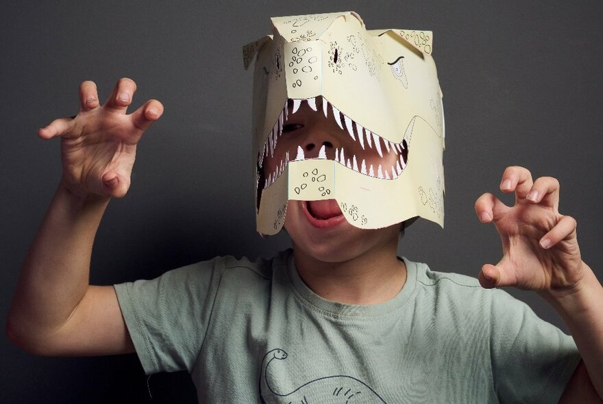 A young child with a hand-made dinosaur mask on, holding his hands up as though they are T. rex claws.