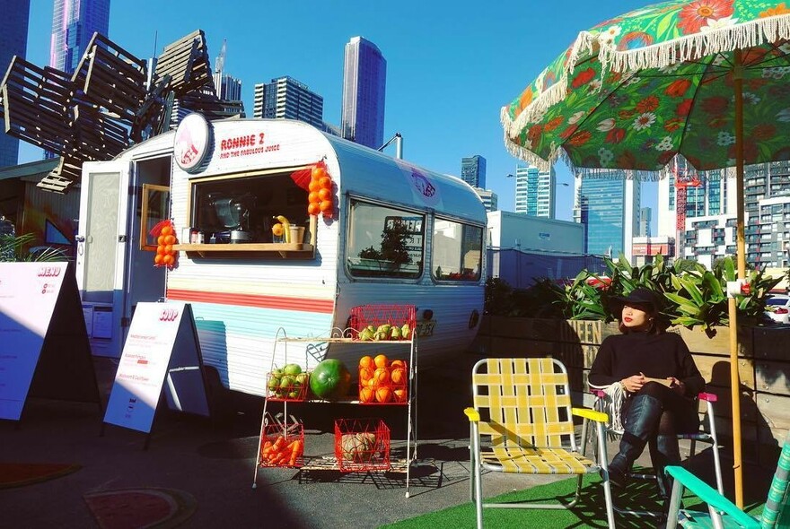 A bright retro caravan, a stand of fruit and a few retro folding chairs and a beach umbrella. 