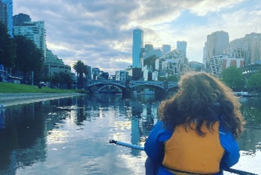 Rear view of a person paddling in a canoe along the Yarra River, with Princes Bridge and city buildings visible in the distance.