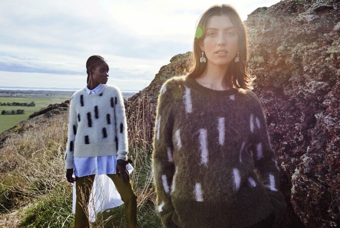 Two models wearing patterned Elk knitwear standing in front of grassy hills in an open field.