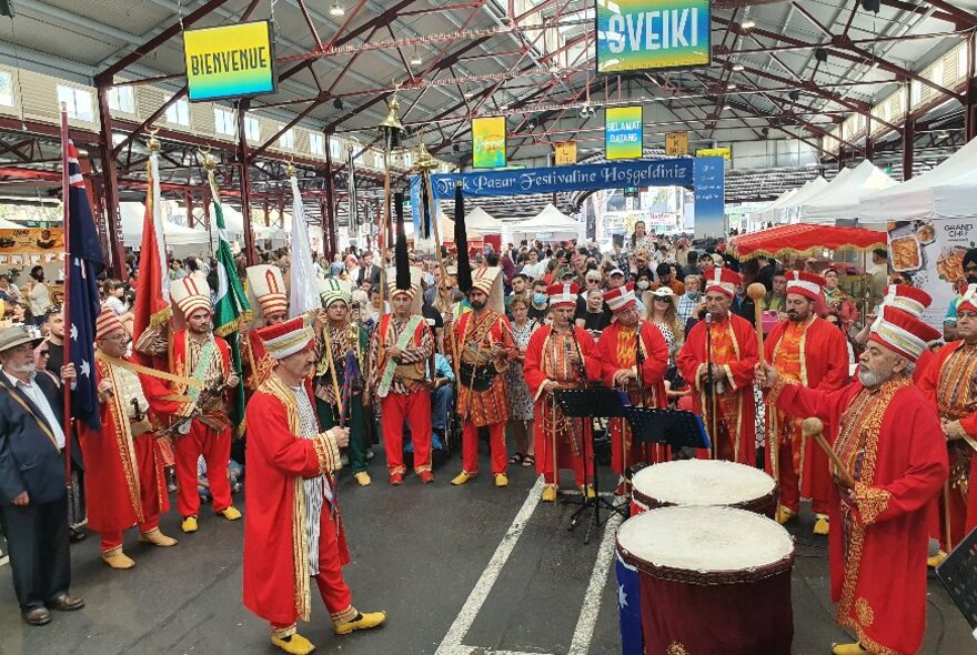 A band of musicians all wearing red cloaks and red pants playing instruments including large drums in an open space under one of the sheds at Queen Victoria Market, with a crowd of onlookers watching.