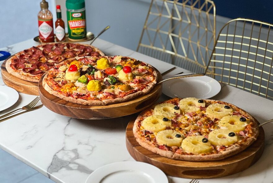 Three pizzas of various kinds on a marble table with wire chairs. 