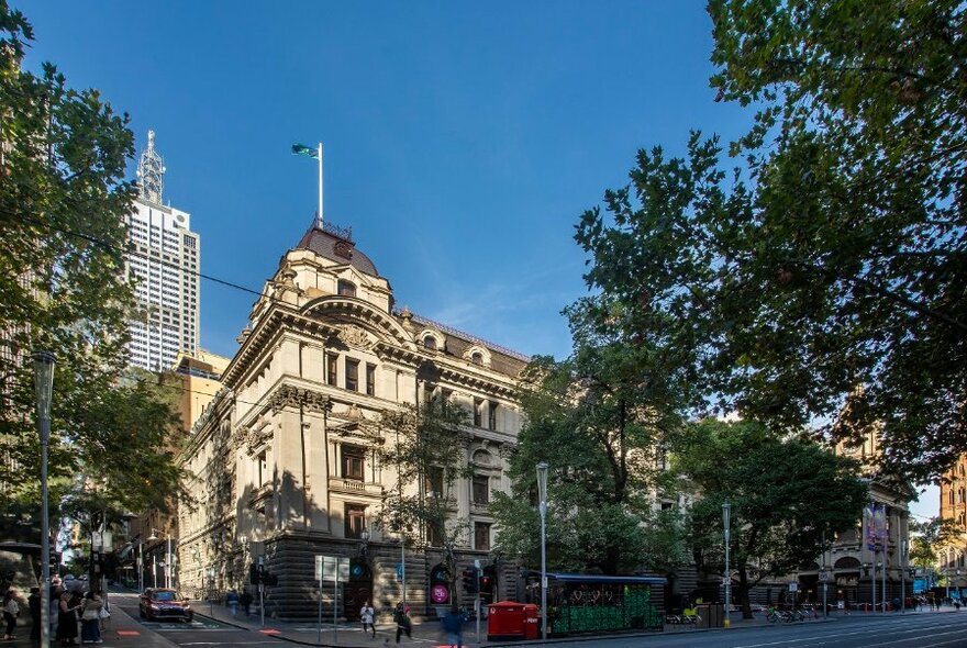 Exterior of Melbourne Town Hall building, with roads in foreground and green trees on either side of the building, blue sky above.