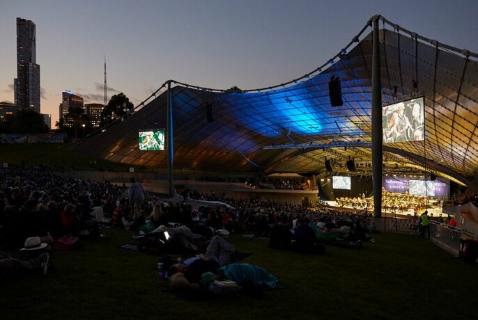 Late evening view of the Sidney Myer Music Bowl structure, with a concert performance taking place on the illuminated stage, and in the foreground people sitting on the lawn watching.