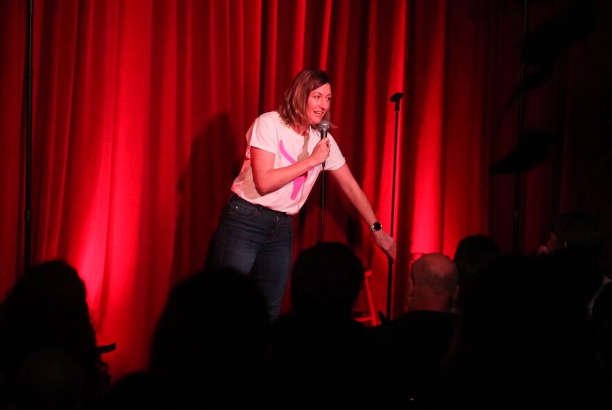 Comedian Celia Pacquola holding a microphone in her hand performing stand-up comedy on a stage, against a red curtain backdrop.