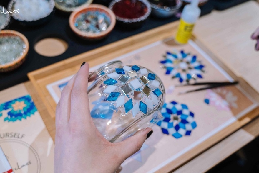 A hand holding a glass bowl decorated with mosaic tiles, tools and dishes in the background.