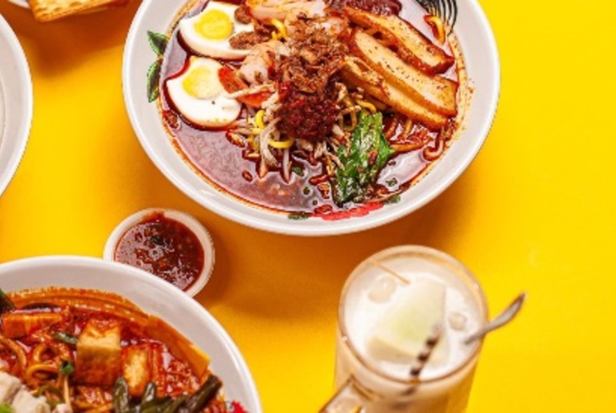 Looking down at two large bowls of curry laksa on a yellow table top, with eggs, bean curd, noodles and broth visible.