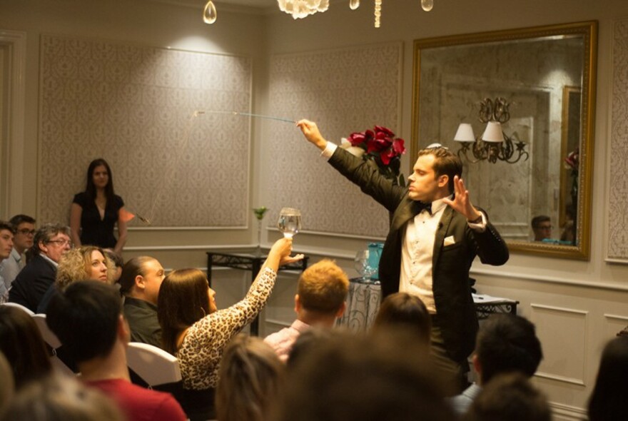 Man wearing a tuxedo and holding a wand, performing in a formal room with seated people, a woman raising her arm in response.
