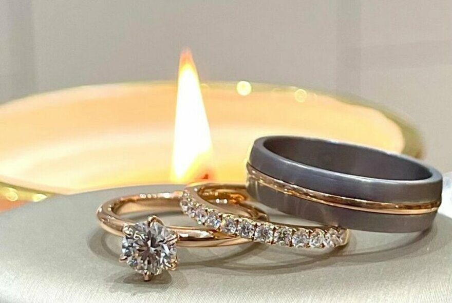 Three rings resting on each other, two with diamonds and a platinum and gold man's ring, a low candle flame in the background.