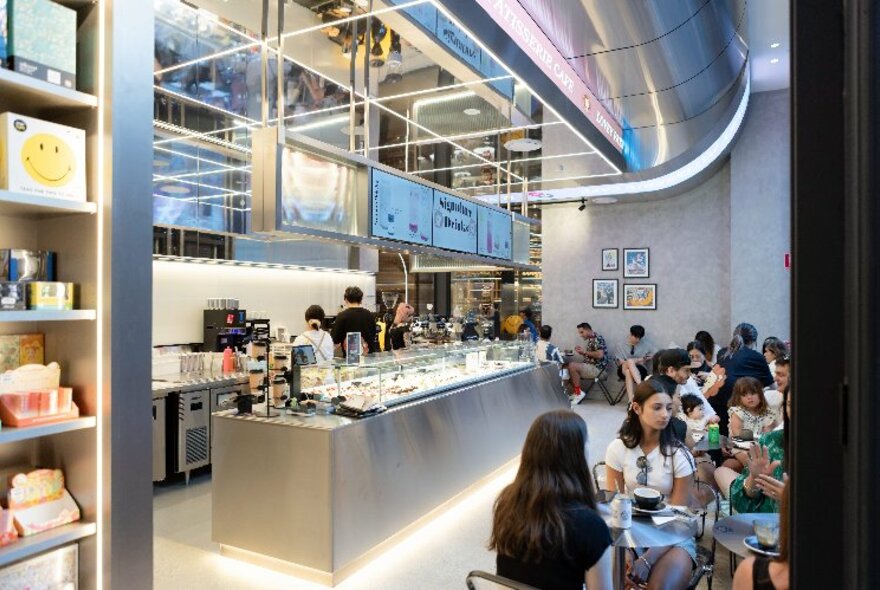 Sleek stainless steel interior of retail cafe space with people seated at tables eating food and drinks, and display areas showing collectible toys and objects.