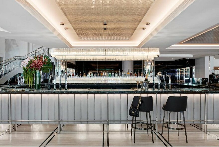 Brightly lit bar, all clean white lines and elegance, with a couple of black stools in front, large flowers at left.