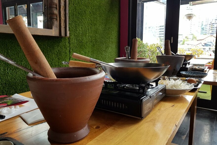 A collection of cookware ready for a Thai cooking workshop, on a timber bench.