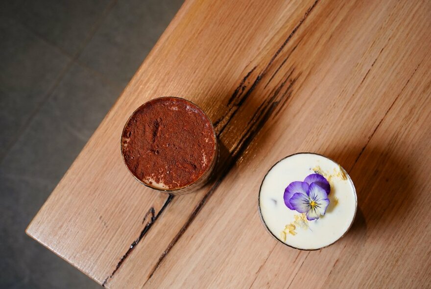 A coffee with a purple nasturtium in its centre next to a hot chocolate with cocoa sprinkled over the top, resting on a wooden table.