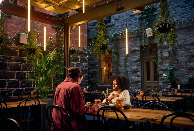 A couple dining in a pub courtyard with bluestone bricks, plants and fairylights.