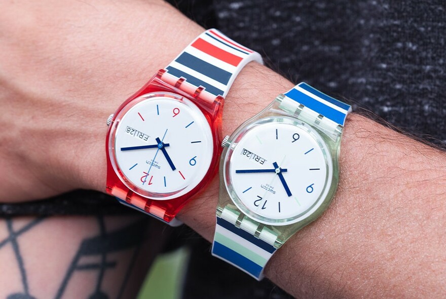 Model's wrist wearing two Swatches, one with blue stripes, the other with red and blue stripes.
