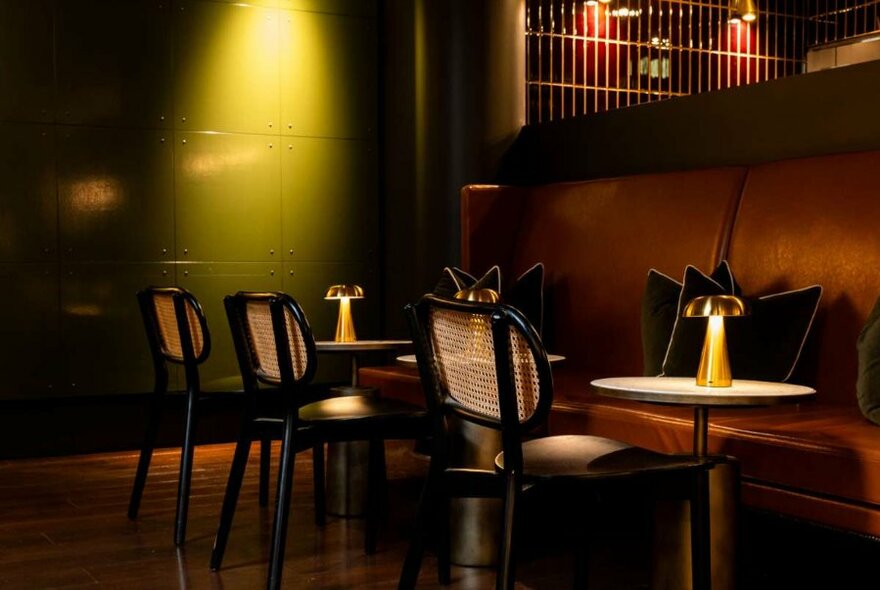 Banquette brown leather seating, small round tables with lamps and rattan dining chairs inside a dimly lit restaurant.