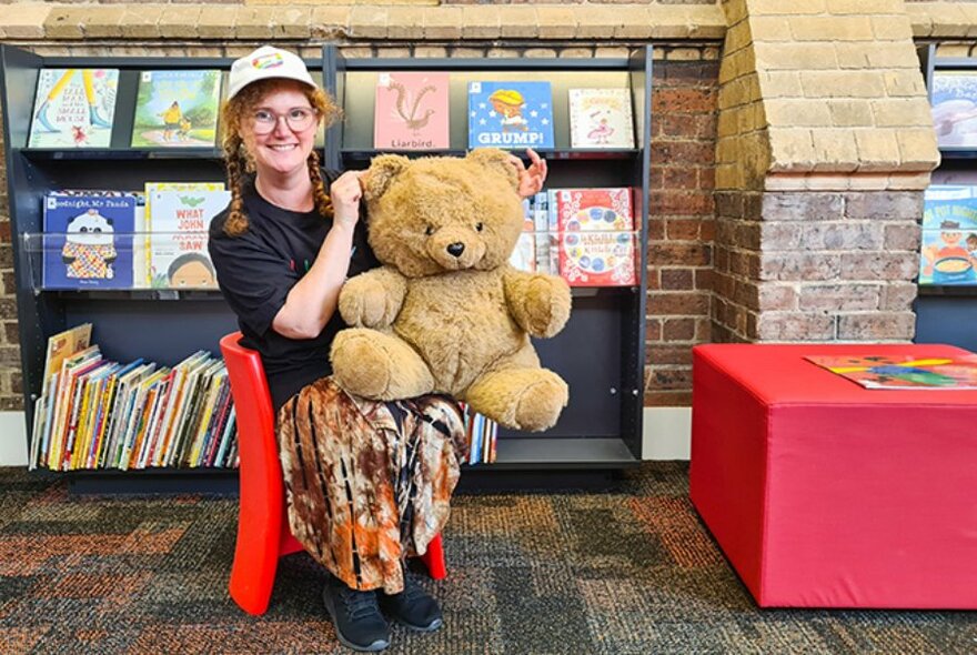 Person seated on a chair in front of a shelf of picture books inside a library, and holding in giant teddy bear on their lap.
