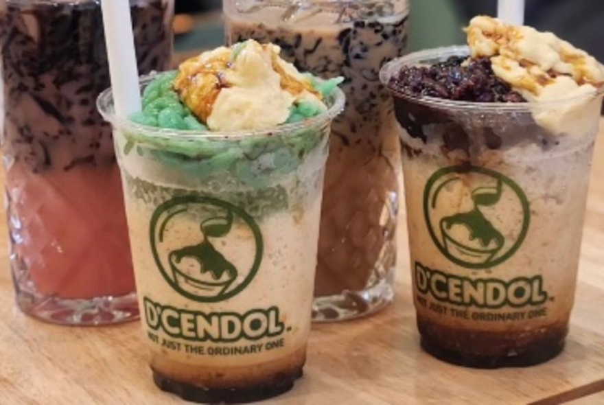 Two plastic takeaway cups of cendol, a loaded dessert drink, each with straws.