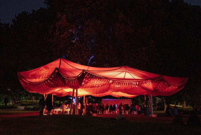 The MPavilion building at night, featuring a red-lit sail design, in a park with people gathering around.