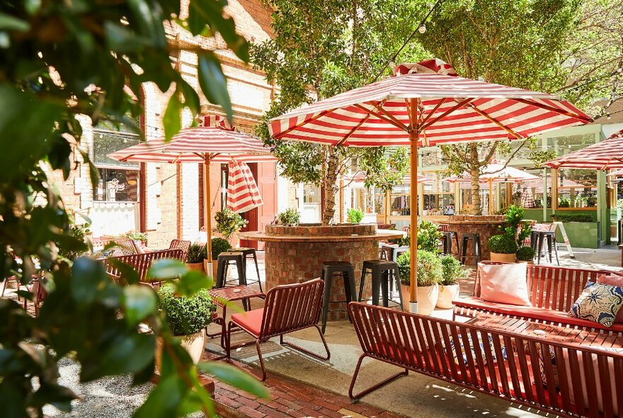 Leafy outdoor courtyard of a restaurant with red and white striped shade umbrellas, and tables and chairs.