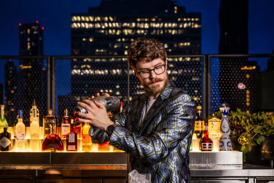 A bartender at an outdoor rooftop bar, wearing a silver jacket and shaking up a drink in a cocktail shaker, illuminated city buildings and the night sky behind him.