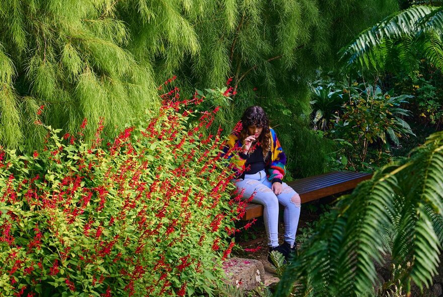 A woman sitting on a garden bench smelling a flower.