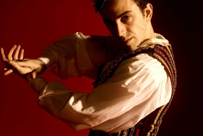 A male dancer with short dark hair, wearing a white shirt with puffy sleeves and a velvet brocade vest, his palms together as if clapping, posed against a red background.