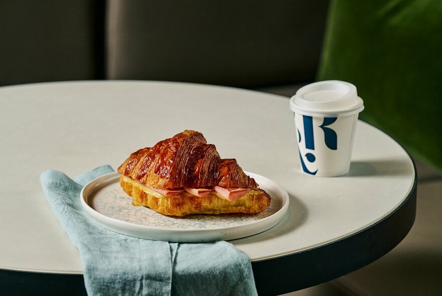 A cafe table with a plate on top of a blue napkin, bearing a filled croissant with a takeaway cup of coffee next to it. 