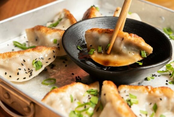 A set of chopsticks dipping a potsticker dumpling into a small bowl of sauce, with more potstickers in a tray underneath.