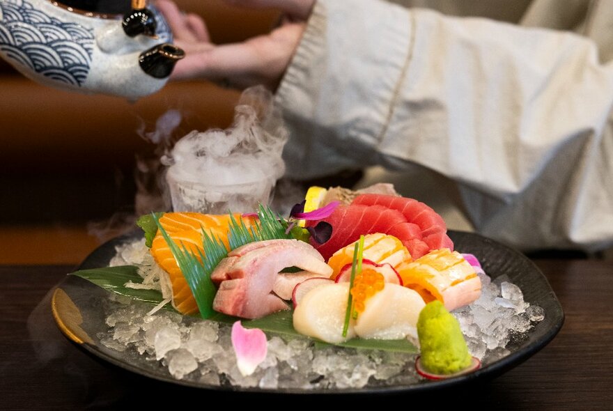 A plate of sashimi with someone pouring tea in the background.
