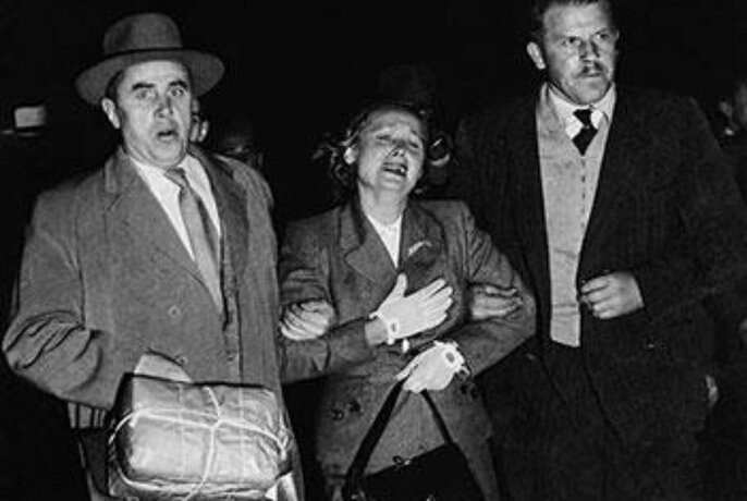 Photo taken at night showing distressed woman flanked by two men in suits holding her arms, circa mid-1950s.