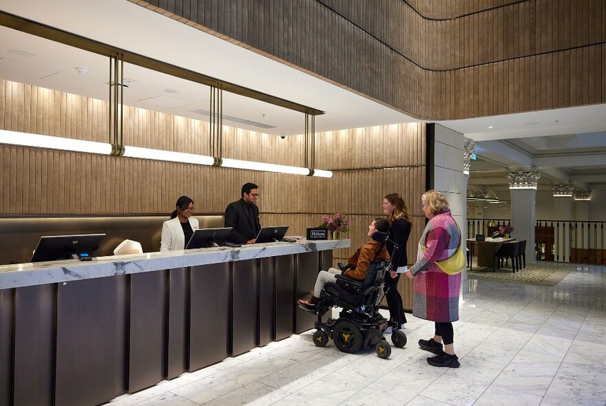 Three people checking in at a hotel lobby, one person is using a wheelchair.