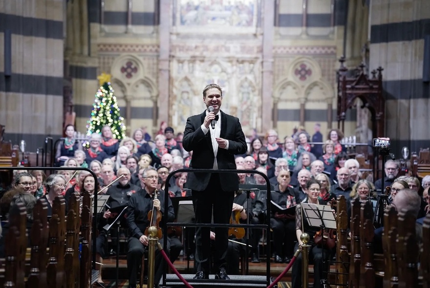 A conductor standing in a church with a choir behind him and a Christmas tree. 