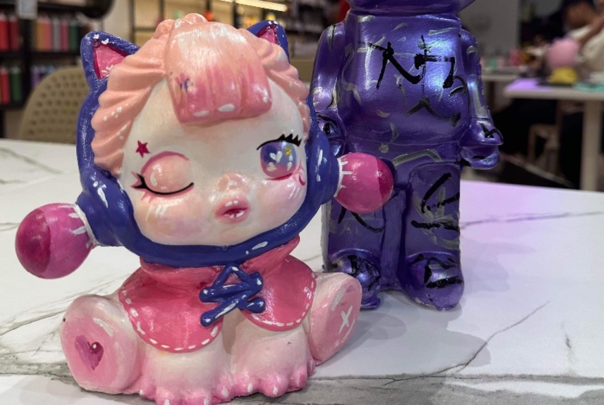 A painted figurine of a cat cartoon character, wearing a purple hood and pink cape, winking.