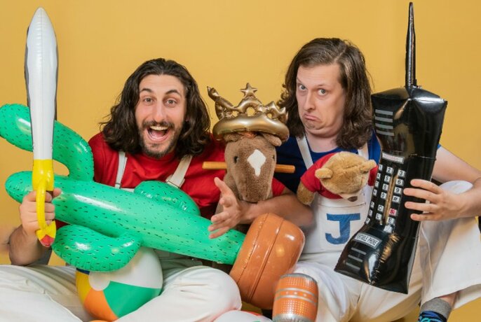 Two men with long hair, holding lots of stuff and pulling funny faces, against a mustard-yellow background.