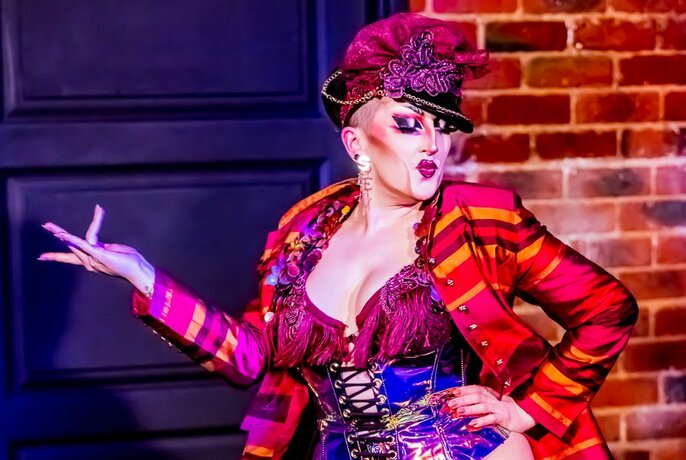 A performer on a stage in front of a brick wall, wearing a multi-coloured burlesque style figure-hugging corset and gaudy jacket and hat, striking an exaggerated pose.