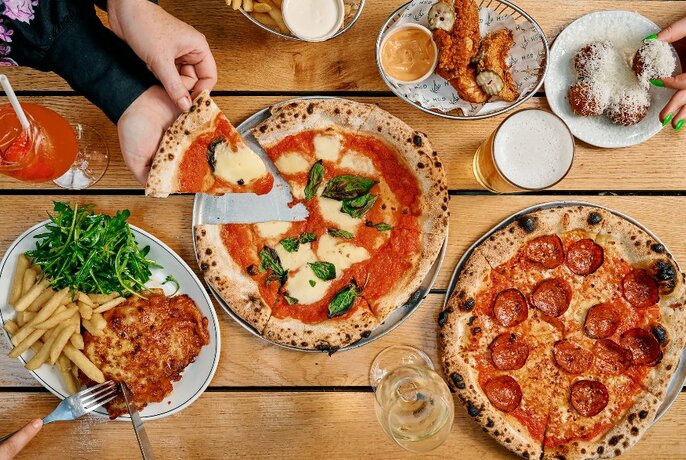 Wooden table seen from above with two pizzas, a parmigiana and chips and a couple of smaller dishes, with hands reaching pizza slice to left and small hands over small dish to right.