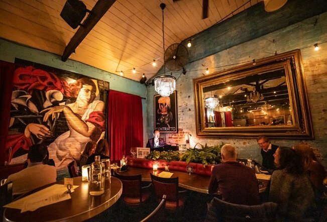People seated in a piano bar at night, with a large mural of a woman on the wall.