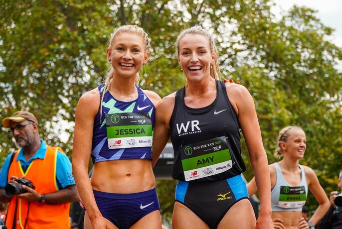 Two women in athletic running shorts and singlets, with their arms around each other's waists, smiling at the camera.