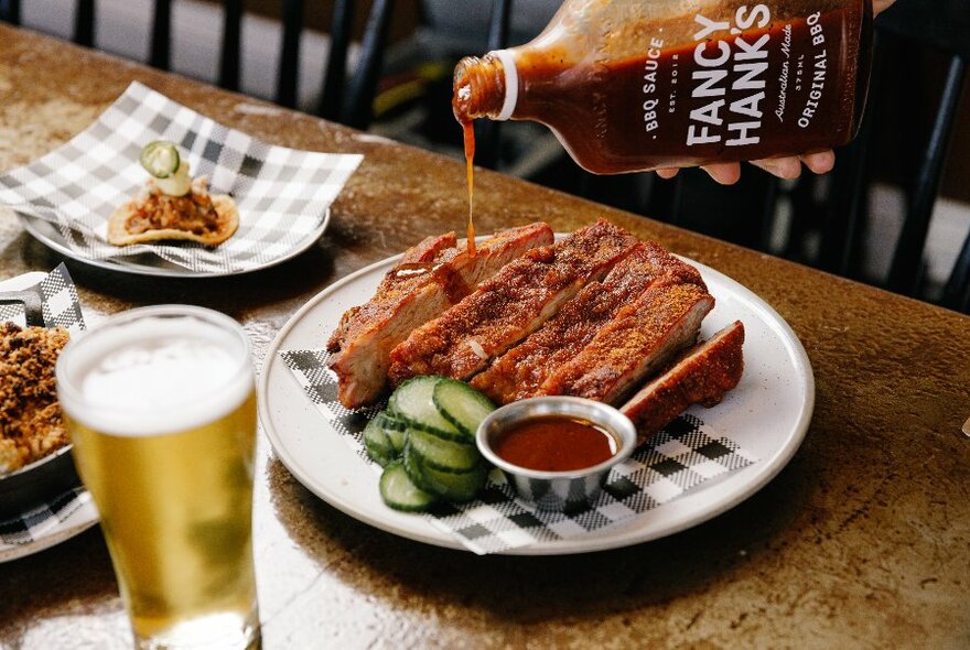 Hand pouring sauce over barbequed meat on a plate, with a glass beer near the plate.