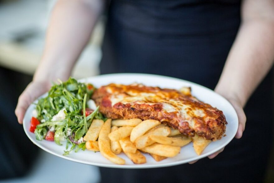 A chicken parma on a plate with chips and salad