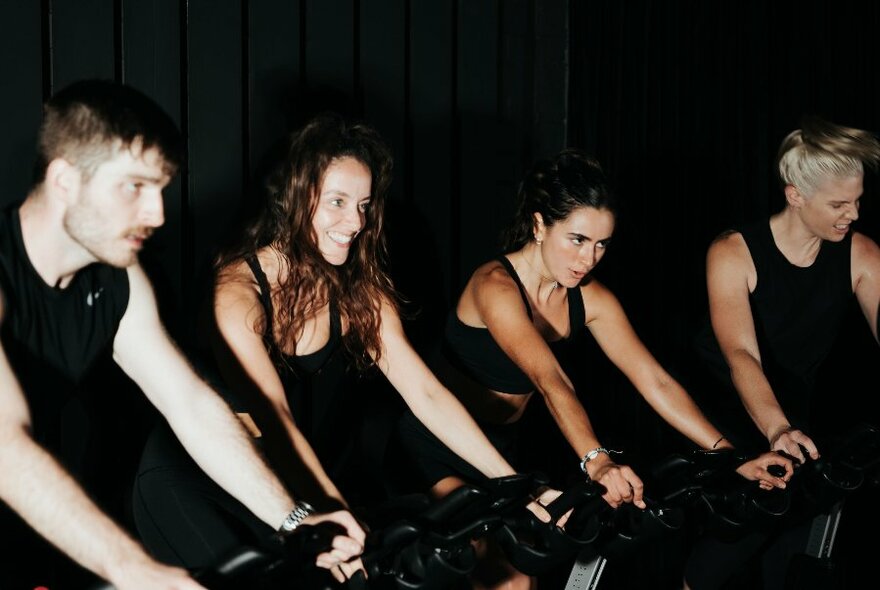 Four people riding stationary bikes in a spin class and wearing black. 