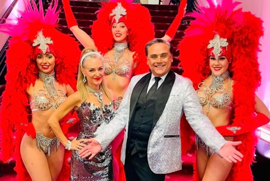 A man in a white jacket poses next to some Vegas showgirls in feathers and diamantés. 