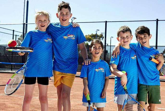 Five children all lined up in matching blue t-shirts on a tennis court. 