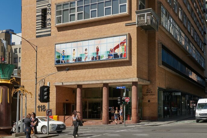 An art installation on the side of a brick office building with people crossing a road outside. 